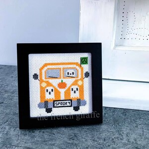 Op de afbeelding: Een ingelijst kruissteekontwerp van een oranje busje met een pompoen aan de voorkant en geesten aan de zijkanten. Het busje heeft het woord "SPOOKY" op de achterkant.