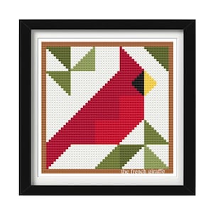 May include: A framed cross-stitch pattern featuring a red cardinal bird with a yellow beak, set against a white background with green and brown geometric shapes. The bottom of the frame includes the text "the french giraffe".