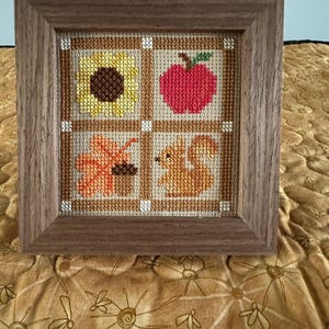 May include: A framed cross-stitch artwork featuring four squares with autumn-themed designs. The squares depict a sunflower, a red apple, a fall leaf, and a squirrel. The frame is made of wood.