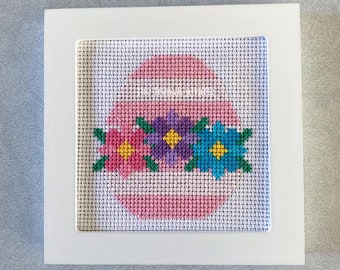 May include: A framed cross-stitch design of a pink egg with three flowers. The flowers are pink, purple, and blue with yellow centers.