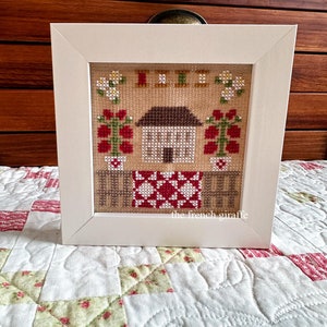 Puede incluir: Arte de punto de cruz enmarcado con una casa, flores y un patrón de edredón en rojo, blanco y marrón. La obra de arte está en un marco blanco y descansa sobre un edredón floral. El texto "the french giraffe" es visible.