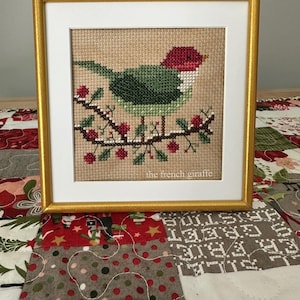 May include: A framed cross-stitch of a green bird with a red head perched on a branch with red berries. The frame is gold with a white mat. The text "the french giraffe" is printed in the bottom right corner of the mat.