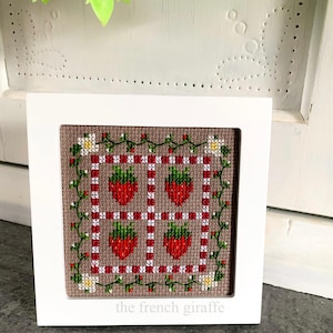 May include: A framed cross-stitch design featuring a brown background with a red and white checkered pattern, green vines, white flowers, and red strawberries. The bottom of the frame has the text "the french giraffe".