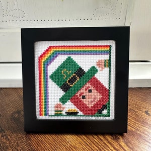 May include: A framed cross-stitch design featuring a cartoon leprechaun holding a pot of gold at the end of a rainbow. The design is titled "The French Giant".
