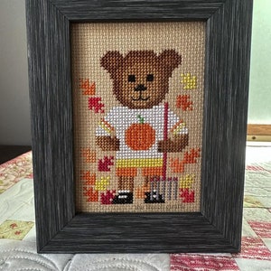 May include: A framed cross-stitch design featuring a brown bear wearing a white shirt with an orange pumpkin design and yellow shorts. The bear is holding a rake and surrounded by orange, yellow, and red leaves. The text "the french giraffe" is at the bottom of the design.