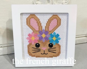 May include: A white framed cross-stitch embroidery of a brown bunny with pink ears and a flower crown. The flowers are pink, purple, and blue.