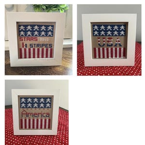 May include: Three framed cross-stitch pieces with patriotic themes. Each features a white frame and a design with blue stars, red and white stripes, and text. The text includes "STARS AND STRIPES", "USA", and "America".