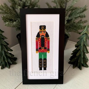 May include: A framed cross-stitch of a traditional nutcracker. The nutcracker is wearing a red and gold uniform with green pants and black boots. The nutcracker has a black beard and mustache and a green hat with a gold star.