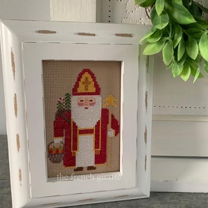Puede incluir: Un bordado de punto de cruz enmarcado en blanco que representa a un tradicional San Nicol&aacute;s con una t&uacute;nica roja, una faja amarilla y un halo dorado. La figura sostiene un bast&oacute;n y una bolsa de regalos. El bordado est&aacute; firmado "the french giraffe".