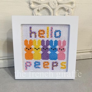 May include: A white framed cross-stitch with the words "Hello Peeps" in colourful letters. The letters are surrounded by five colourful bunny-shaped figures.