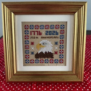 Op de afbeelding: Ingelijst kruissteekkunstwerk met een gouden lijst. Het ontwerp toont een Amerikaanse zeearend, de tekst "1776-2026 250th Anniversary" en rode, witte en blauwe accenten. Het kunstwerk staat op een beige achtergrond.