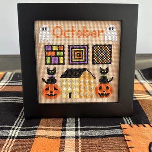 May include: A framed cross-stitch design featuring a house, pumpkins, cats, and ghosts. The design is titled "October" and includes a variety of colorful patchwork squares.
