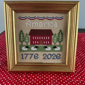 May include: Framed cross-stitch art featuring a red building, green trees, and sheep on a green field. The word "America" is stitched above the building, with the years "1776 2026" below. The frame is gold.