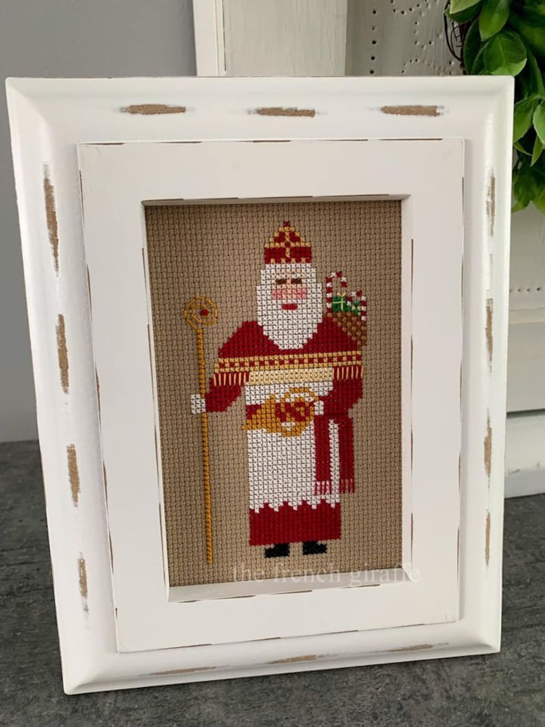 K&ouml;nnte beinhalten: Ein wei&szlig; gerahmter Kreuzstich eines roten und wei&szlig;en Weihnachtsmanns, der einen Korb mit Geschenken h&auml;lt. Der Kreuzstich ist auf beigefarbenem Hintergrund und der Rahmen hat eine wei&szlig;e, abgenutzte Oberfl&auml;che.