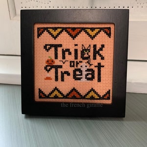 Könnte beinhalten: Ein quadratischer schwarzer Holzrahmen mit einer Kreuzstichstickerei auf pfirsichfarbenem Stoff. Das Design zeigt den schwarzen Schriftzug Trick or Treat, einen kleinen orangefarbenen Kürbis, ein weißes Gespenst und eine schwarze Katze.