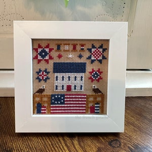 May include: A framed cross-stitch design featuring a white house with red and blue accents, a red, white, and blue American flag, and two birds. The design is surrounded by stars and other patriotic symbols. The frame is white and the text "the french giraffe" is visible at the bottom.