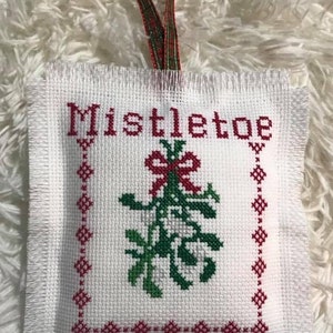 May include: White fabric ornament with a red and green embroidered mistletoe design and the word "Mistletoe" in red lettering. The ornament has a frayed edge and a red and green ribbon for hanging.