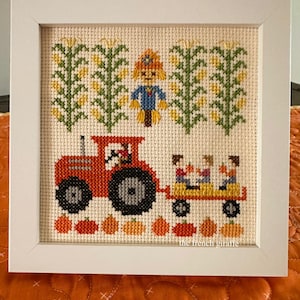 May include: Cross-stitch picture in a white frame showing an autumn scene. A red tractor pulls a wagon with children. Corn stalks and a scarecrow are in the background. Pumpkins are at the bottom. The text "the french giraffe" is in the bottom right corner.