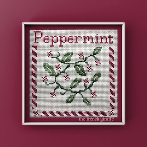 May include: A white framed cross-stitch design with a red and white striped border. The design features a green peppermint sprig with red berries and the word "Peppermint" in red lettering. The bottom right corner of the frame has the text "the french giraffe".