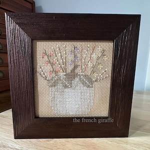 May include: A framed cross-stitch piece depicting a white ceramic vase filled with colorful flowers. The vase is sitting on a brown burlap background. The frame is dark brown wood with a simple design. The text "the french giraffe" is printed at the bottom of the frame.