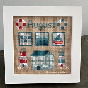 May include: A framed cross-stitch design with a nautical theme. The design features a lighthouse, a house, a sailboat, a life preserver, and a window. The text "August" is stitched in the center of the design.