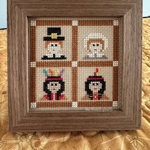 May include: A framed cross-stitch artwork featuring four squares with Thanksgiving-themed figures. The figures include a pilgrim, a woman in a bonnet, and two Native American figures. The frame is made of wood.