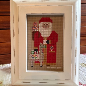 May include: A white framed cross-stitch featuring a Santa Claus holding a bag of gifts and a llama with presents. The cross-stitch is signed "the french giraffe".