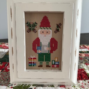 Puede incluir: Un bordado de punto de cruz enmarcado de un gnomo con gorro y chaqueta rojos, pantalones verdes y un regalo. La obra de arte está en un marco blanco desgastado. El texto "the french giraffe" está en la parte inferior.