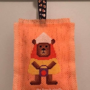 May include: A cross-stitch ornament featuring a brown bear wearing a yellow, orange, and white candy corn costume. The bear is holding a small orange bucket. The ornament is on a peach-colored background and has a frayed edge. The text "the french giraffe" is at the bottom of the ornament.