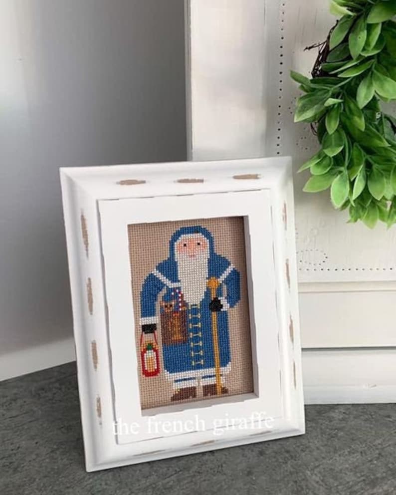 May include: A framed cross-stitch of Santa Claus in a blue coat, holding a lantern and a walking stick. The artwork is set against a tan background and framed in a distressed white wooden frame. The text "the french giraffe" is at the bottom.