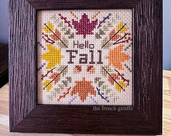 May include: A framed cross-stitch with a brown wooden frame. The cross-stitch features a design of leaves and the words "Hello Fall" in a white font.