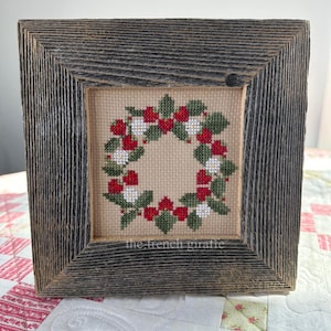 May include: A framed cross-stitch design of a red and white berry wreath with green leaves. The frame is made of rustic wood and has a weathered gray finish. The text "the french giraffe" is visible at the bottom of the design.