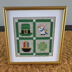 May include: A framed cross-stitch design with four squares. The top left square shows a green leprechaun hat, the top right square shows a white unicorn with a rainbow mane, the bottom left square shows a pot of gold with a rainbow, and the bottom right square shows a green shamrock. The frame is gold and the cross-stitch is on a beige linen fabric. The text "the french giraffe" is at the bottom of the design.