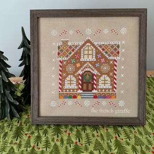 May include: A cross-stitch pattern of a gingerbread house with candy cane trim, candy decorations, and a wreath on the door. The pattern is framed in a brown wood frame and the text "the french giraffe" is in the bottom right corner.