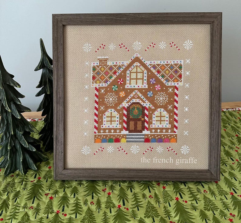May include: A cross-stitch pattern of a gingerbread house with candy cane trim, candy decorations, and a wreath on the door. The pattern is framed in a brown wood frame and the text "the french giraffe" is in the bottom right corner.