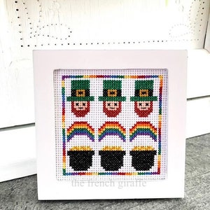 Peut inclure: Un cadre blanc avec un motif de point de croix représentant trois lutins portant des chapeaux verts et tenant des pots d'or au bout d'arcs-en-ciel. Le texte "the french giraffe" est en bas du cadre.