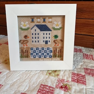 Puede incluir: Un diseño de punto de cruz enmarcado que presenta una casa blanca con un techo azul, dos patos y flores blancas. El punto de cruz está firmado "the french giraffe".