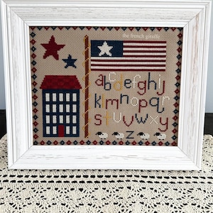 May include: A framed cross-stitch sampler with a red, white, and blue American flag, a red house with white windows, and the alphabet in blue letters. The sampler is on a white crocheted doily.