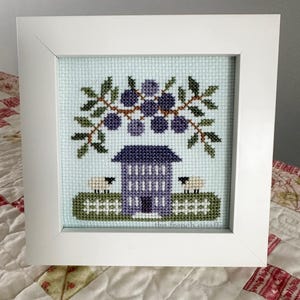 May include: A framed cross-stitch design featuring a purple house with a white door, two white sheep, and a green bush with purple berries. The text "The French Giraffe" is at the bottom of the design.