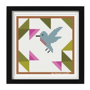 Peut inclure: Un motif de point de croix représentant un colibri bleu avec des fleurs roses et des feuilles vertes dans un design géométrique. Le motif est encadré dans un cadre noir avec un passe-partout marron. Le texte "the french giraffe" est en bas du motif.