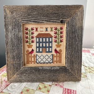 May include: A framed cross-stitch piece depicting a blue house with red cardinals and green bushes. The frame is made of rustic wood and has a weathered gray finish. The text "the french giraffe" is visible at the bottom of the image.