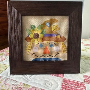 May include: A framed cross-stitch of a smiling scarecrow with a straw hat, a sunflower, and a blue and white checked pattern. The frame is brown wood.