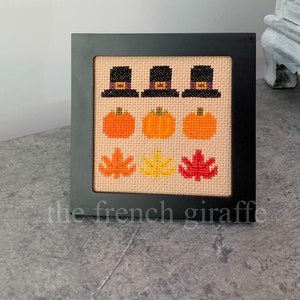 May include: A framed cross-stitch design featuring three pilgrim hats, three pumpkins, and three autumn leaves in shades of orange, yellow, and red.