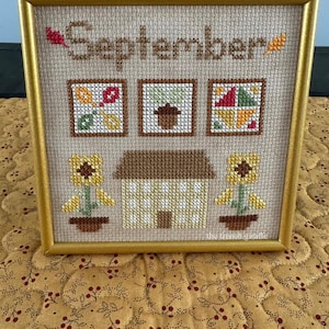 May include: A framed cross-stitch design featuring the word "September" with autumnal motifs including sunflowers, a house, and a patchwork quilt. The design is stitched on beige fabric and framed in a gold frame.