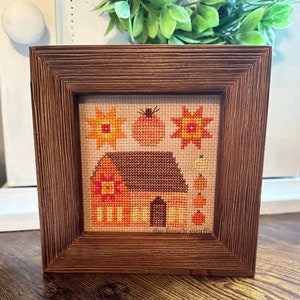 May include: A framed cross-stitch design featuring a house with a pumpkin and sunflowers. The design is in shades of orange, yellow, and brown. The frame is brown wood.