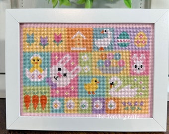 May include: A framed cross-stitch design featuring a patchwork of colorful squares with Easter-themed images, including a bunny, chicks, eggs, flowers, and a house. The text "the french giraffe" is visible in the bottom right corner.