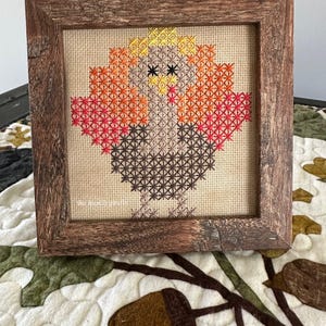 May include: A framed cross-stitch artwork featuring a colorful bird design. The bird has a yellow, orange, and red head and wings, with a brown body and legs. The artwork is set in a rustic, square wooden frame.