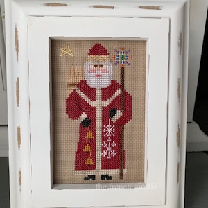 May include: A framed cross-stitch of Santa Claus in a red coat with a white beard and a golden halo. He is holding a staff with a star on top and a bag of gifts on his back. The cross-stitch is on a beige background with snowflakes and bells. The frame is white with a distressed finish. The text "the french giraffe" is at the bottom of the cross-stitch.