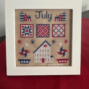 May include: A framed cross-stitch design featuring a red, white, and blue color scheme. The design includes the word "July", a house, pinwheels, and stars. The cross-stitch is on a beige fabric.