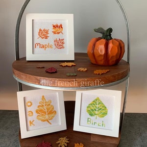 May include: Three framed cross-stitch embroidery pieces featuring different types of leaves. The top frame shows three maple leaves in shades of orange and red. The bottom two frames show a single oak leaf and a single birch leaf. The frames are white and the background is a wooden tiered stand.